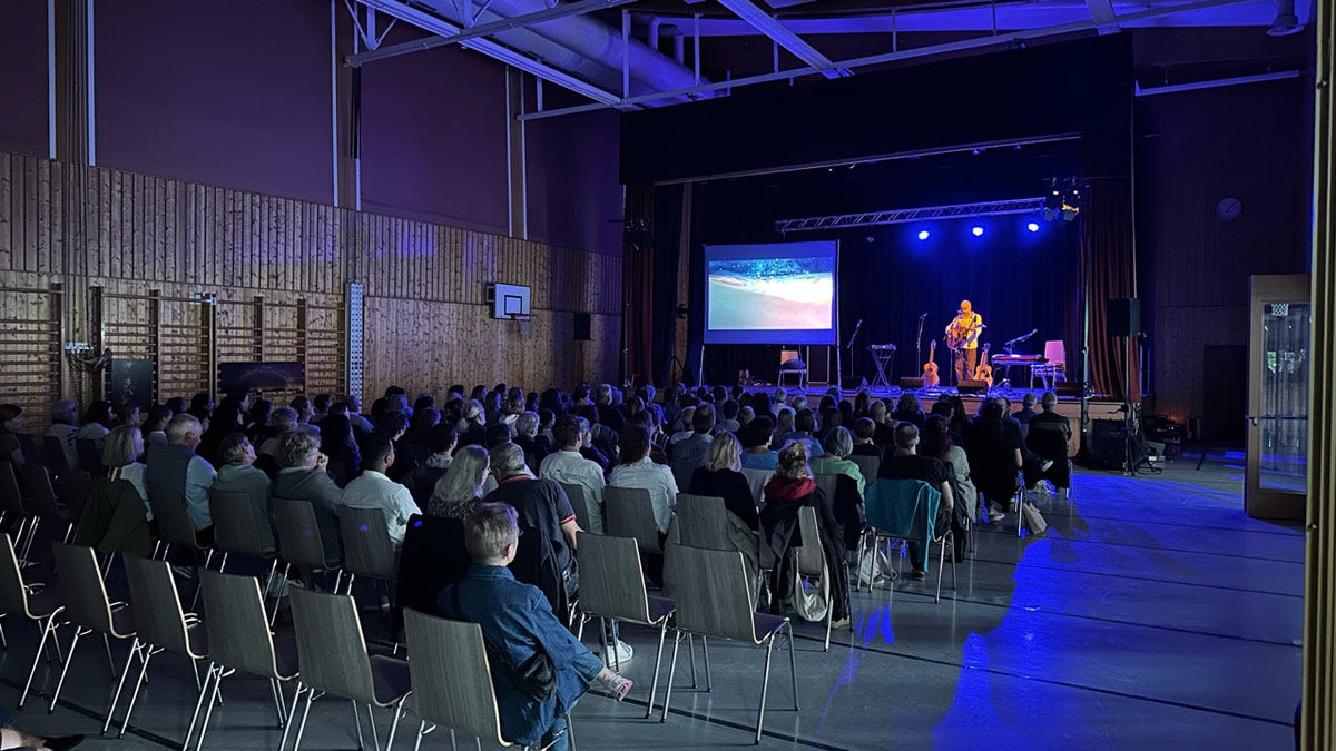 Public assis face à une scène éclairée en bleu, avec un musicien et des images projetées dans la salle polyvalente d’Arbaz.