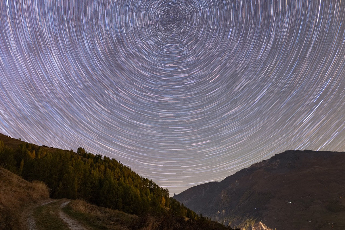 Filés d’étoiles en arcs concentriques au-dessus d’une vallée de montagne, avec un chemin et une forêt au premier plan aux Haudères.