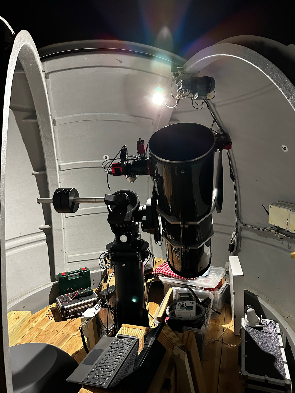Intérieur du dôme blanc avec un grand télescope noir sur sa monture, un ordinateur au premier plan et l’ouverture vers le ciel nocturne.