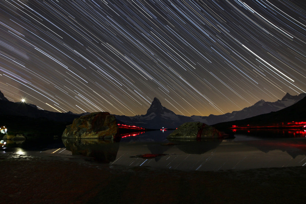 Filés d’étoiles obliques au-dessus du Cervin, avec des rochers et le lac du Stellisee au premier plan.