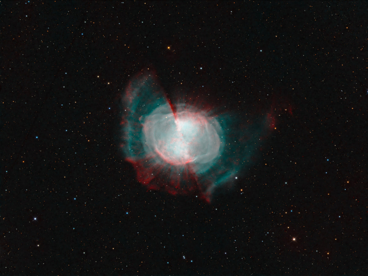 Nébuleuse de l’Haltère au centre clair, entourée de lobes bleutés et rougeâtres dans un champ d’étoiles.