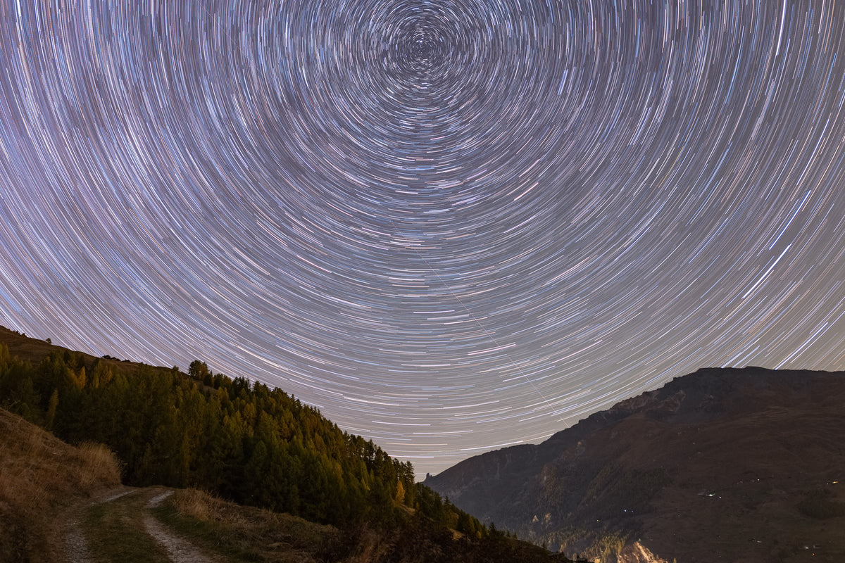 Arcs concentriques de filés d’étoiles dans un ciel violet, au-dessus de montagnes et de silhouettes d’arbres.