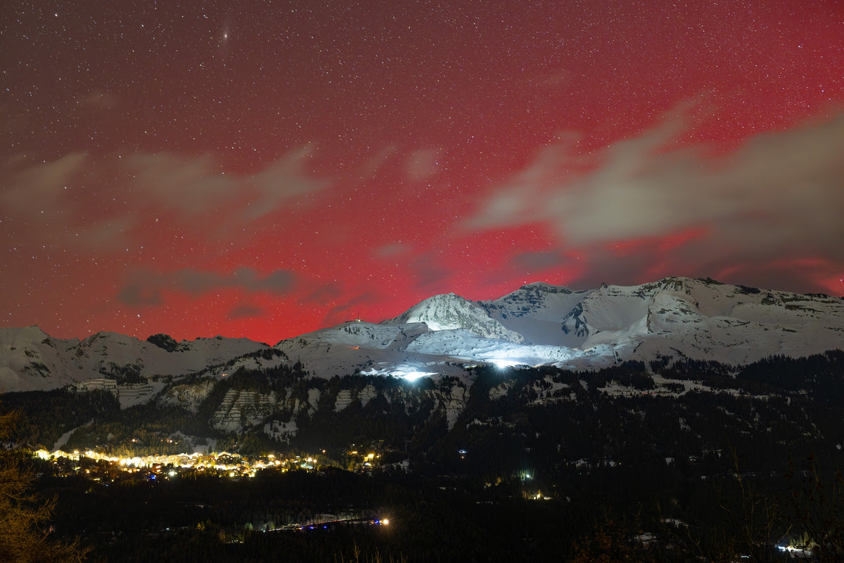 Large ciel rouge d’aurore au-dessus d’une chaîne enneigée et des lumières d’Anzère, avec des nuages au premier plan.