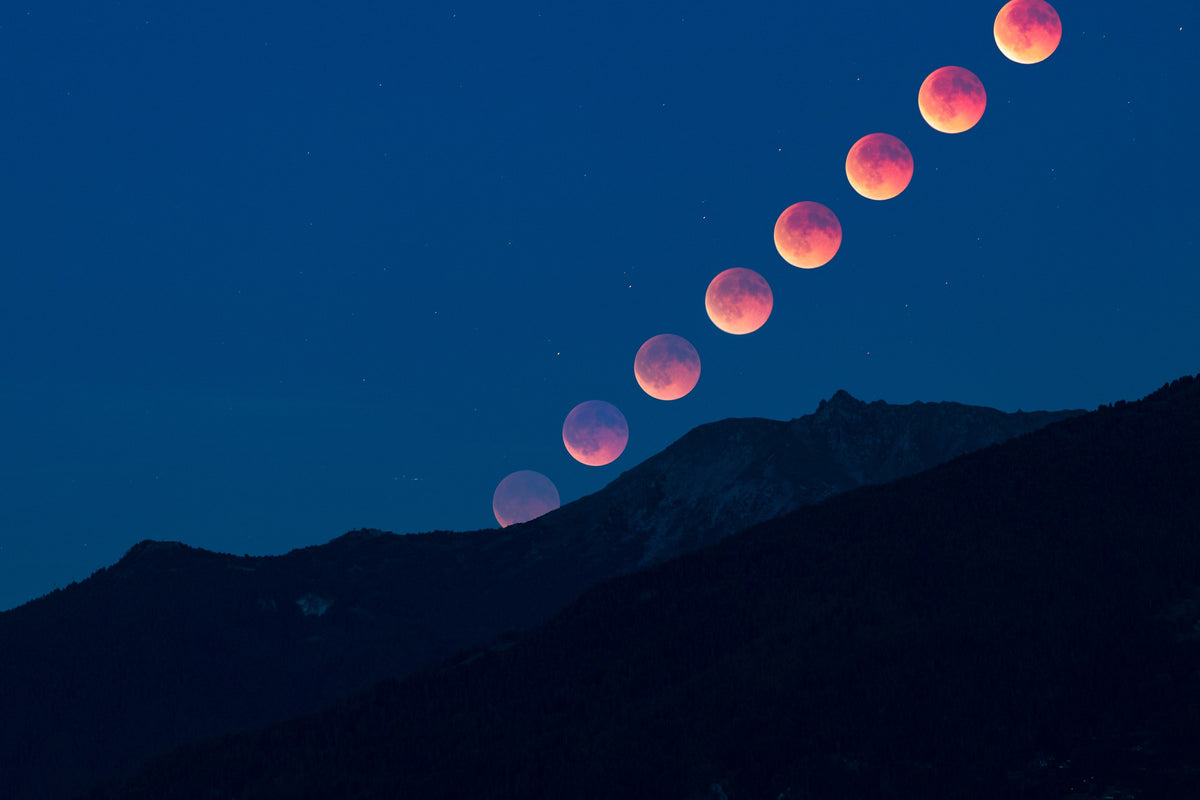 Disques successifs de la Lune rouge en diagonale au-dessus d’une crête sombre, sur un ciel bleu.