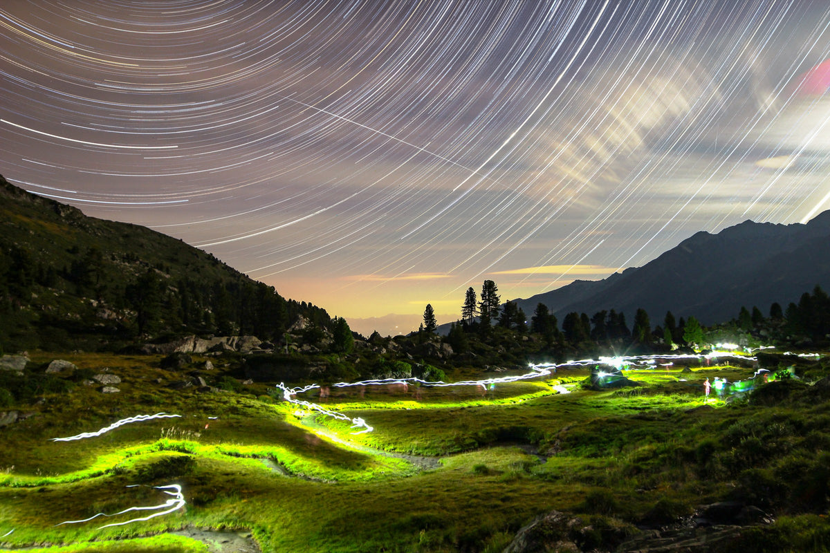 Filés d’étoiles au-dessus des montagnes, avec des lignes lumineuses jaunes et vertes dans le terrain rocheux.
