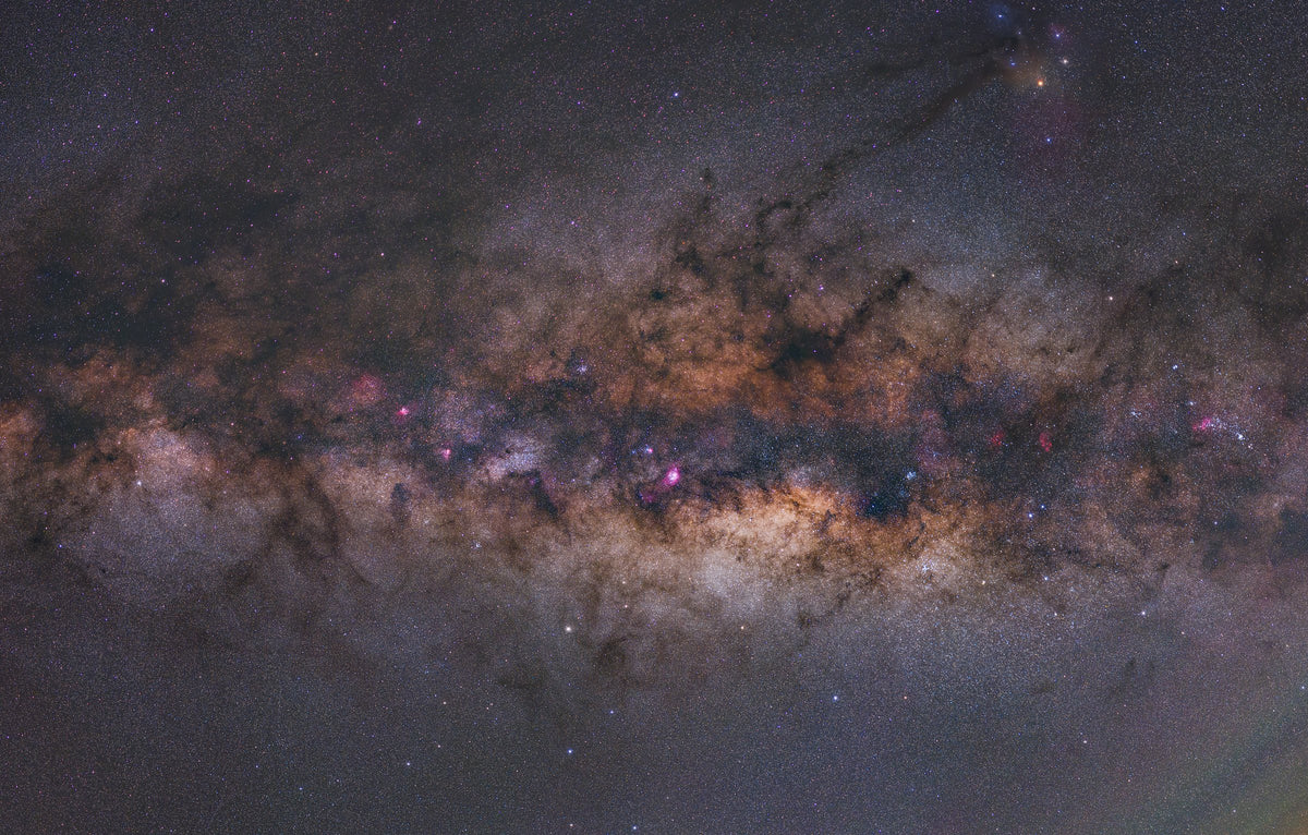 Large champ du centre de la Voie lactée, aux teintes dorées et rosées, traversé de nuages sombres.