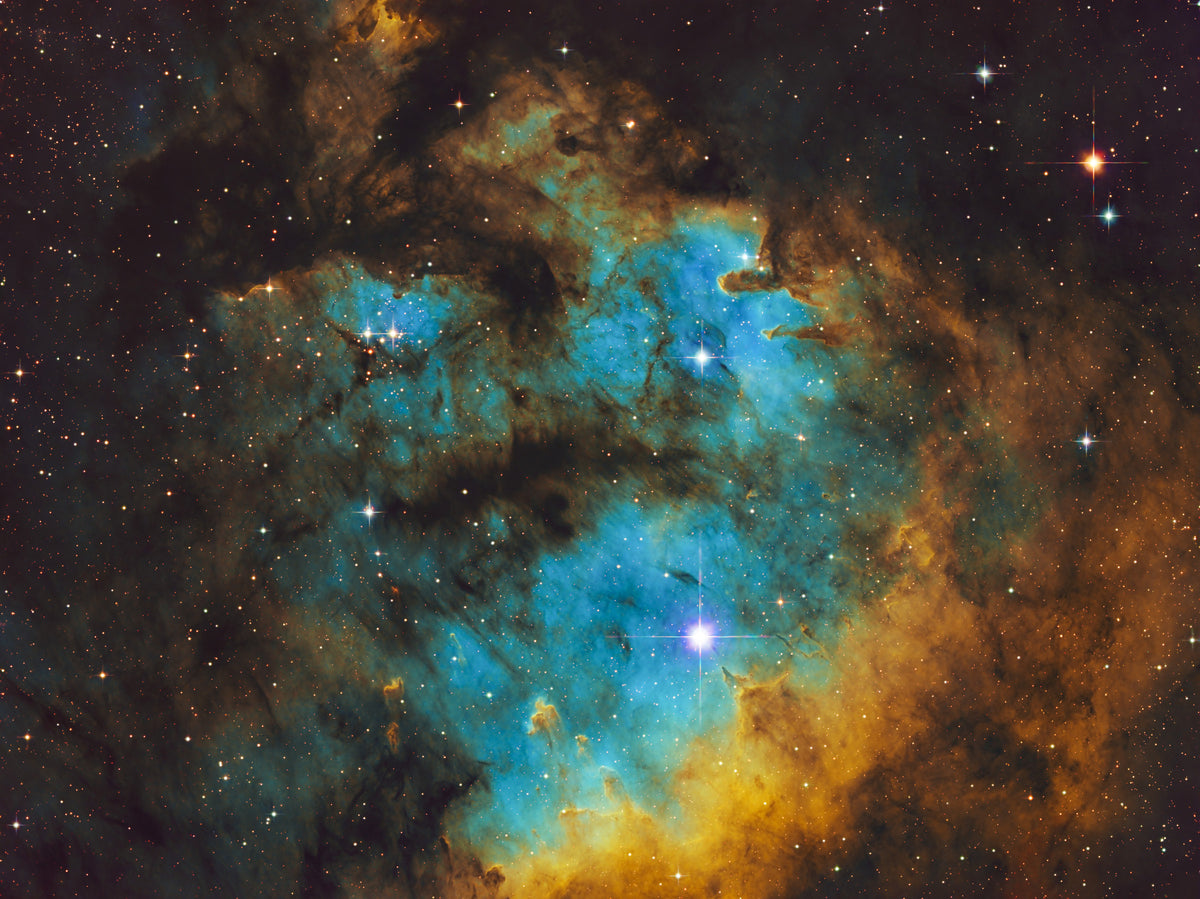 Nébuleuse turquoise et dorée de NGC 7822, aux bords irréguliers et aux structures sombres parsemées d’étoiles.