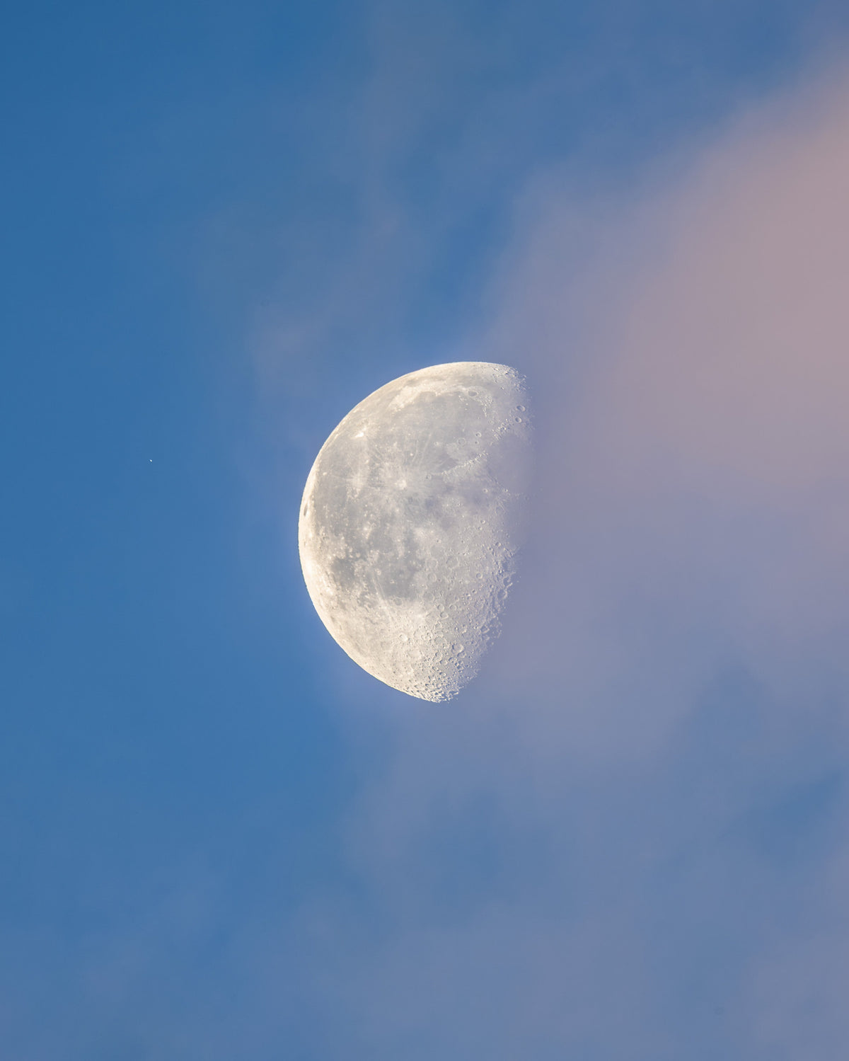 Lune partiellement éclairée dans un ciel bleu, à côté de voiles nuageux rosés.