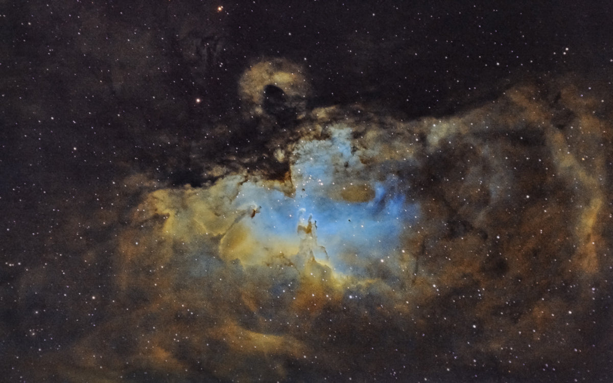 Nébuleuse de l’Aigle aux nuages bleutés et dorés, traversés par des structures sombres et ponctués d’étoiles.