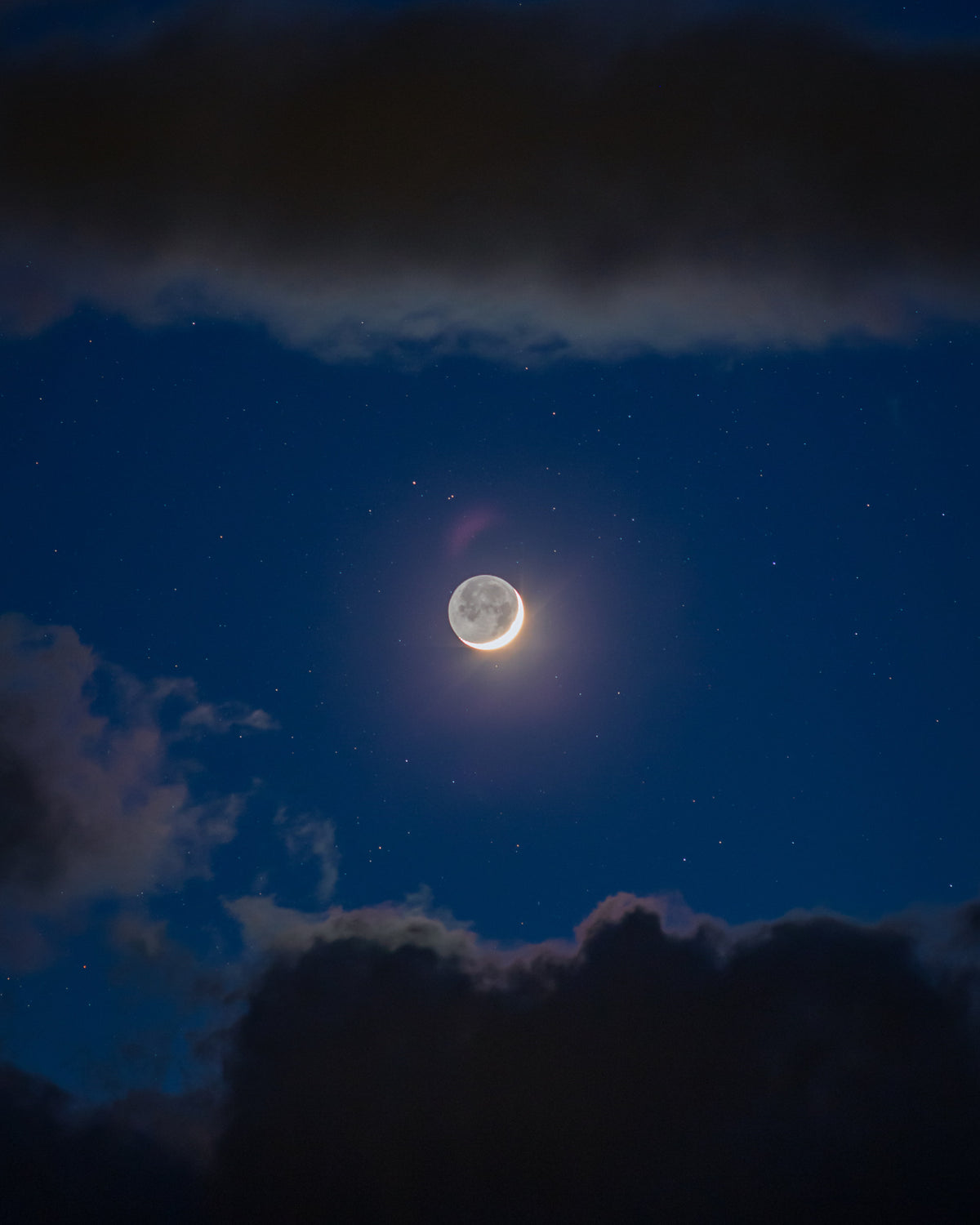Jeune Lune au milieu d’un ciel bleu, encadrée au-dessus et au-dessous par de gros nuages sombres.