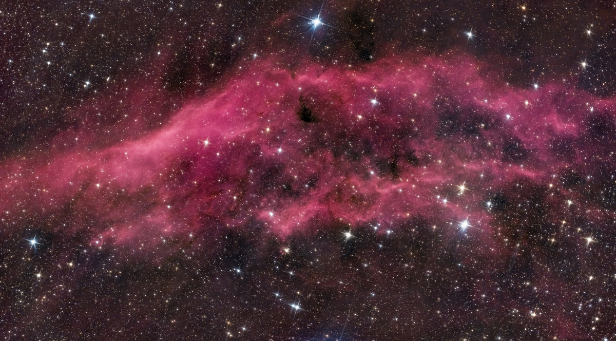 Longue nébuleuse rose-rouge aux contours irréguliers, bordée de zones sombres et d’étoiles.