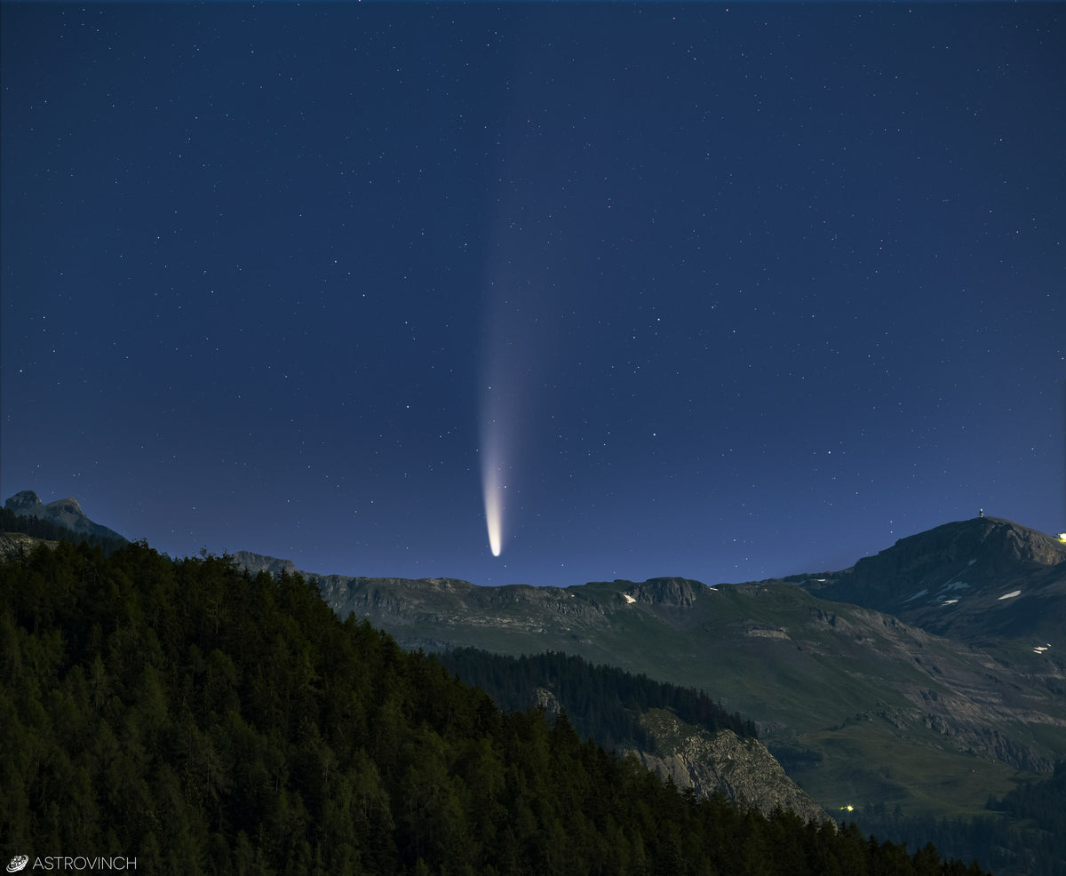 Comète NEOWISE à la queue presque verticale dans un ciel bleu sombre, au-dessus de collines boisées.