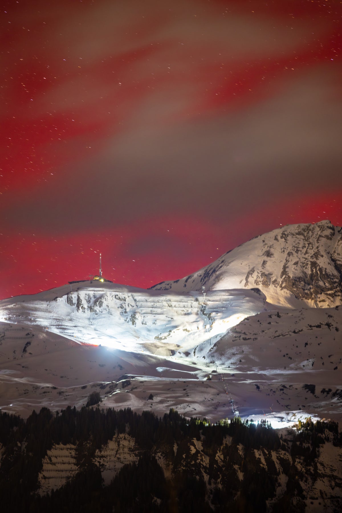 Ciel rouge parcouru de voiles d’aurore au-dessus d’Anzère, des montagnes enneigées et du village éclairé.