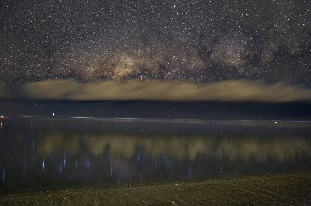 Voie lactée presque horizontale au-dessus de l’océan, avec des reflets lumineux dans l’eau au bord d’une plage.