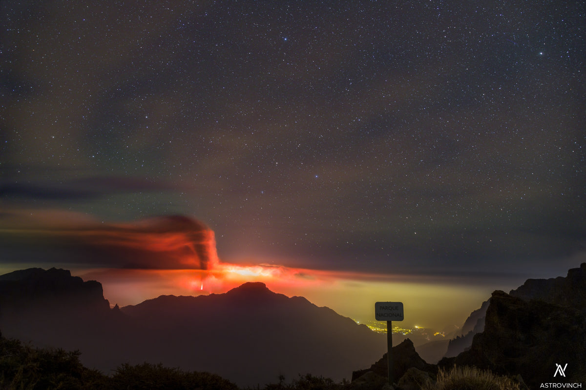 Fontaine et lueur rouges d’un volcan en éruption, au-dessus de collines sombres sous des nuages et des étoiles.