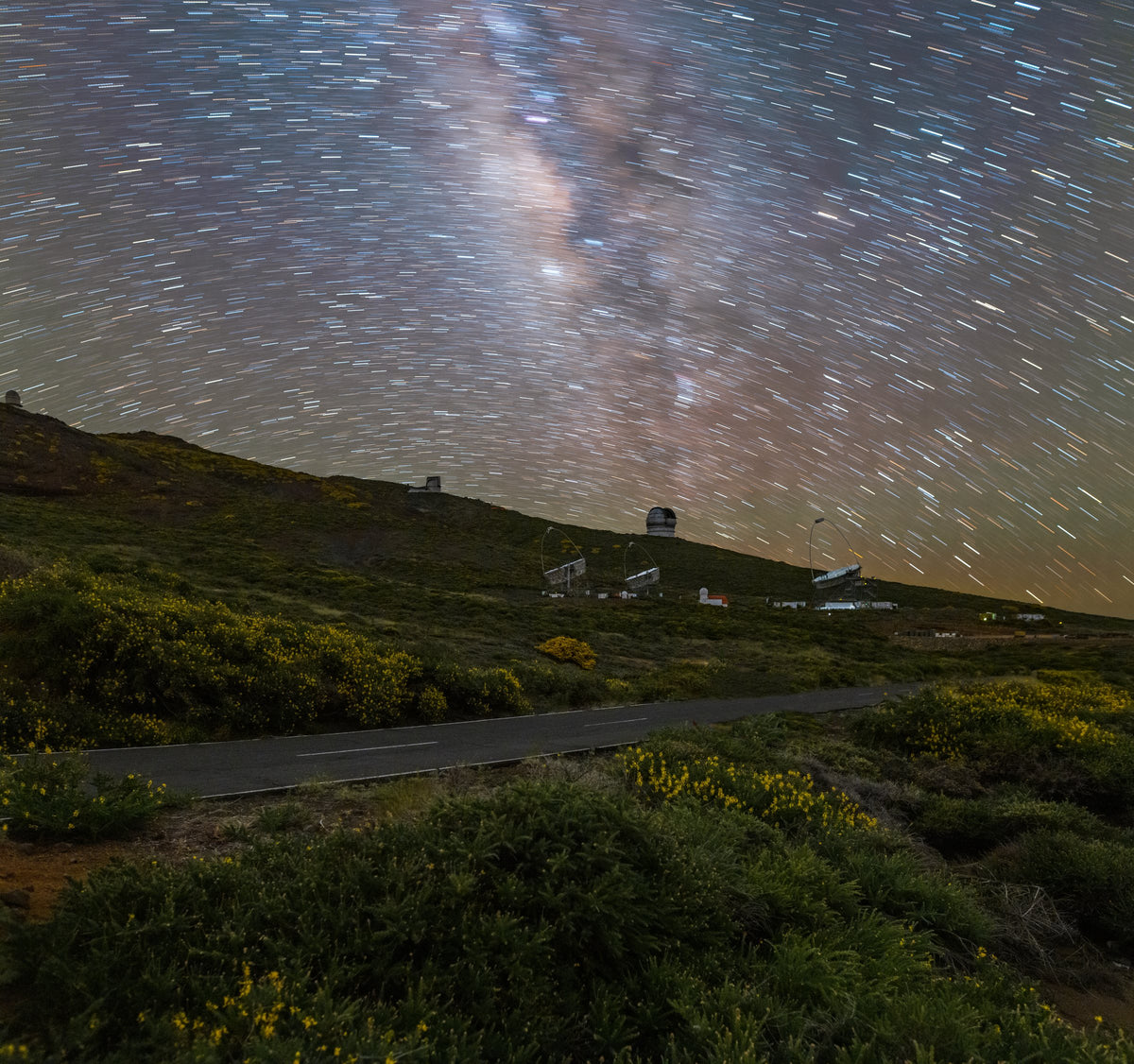 Courts filés d’étoiles et Voie lactée au-dessus de coupoles et de télescopes, sur une pente traversée par une route.