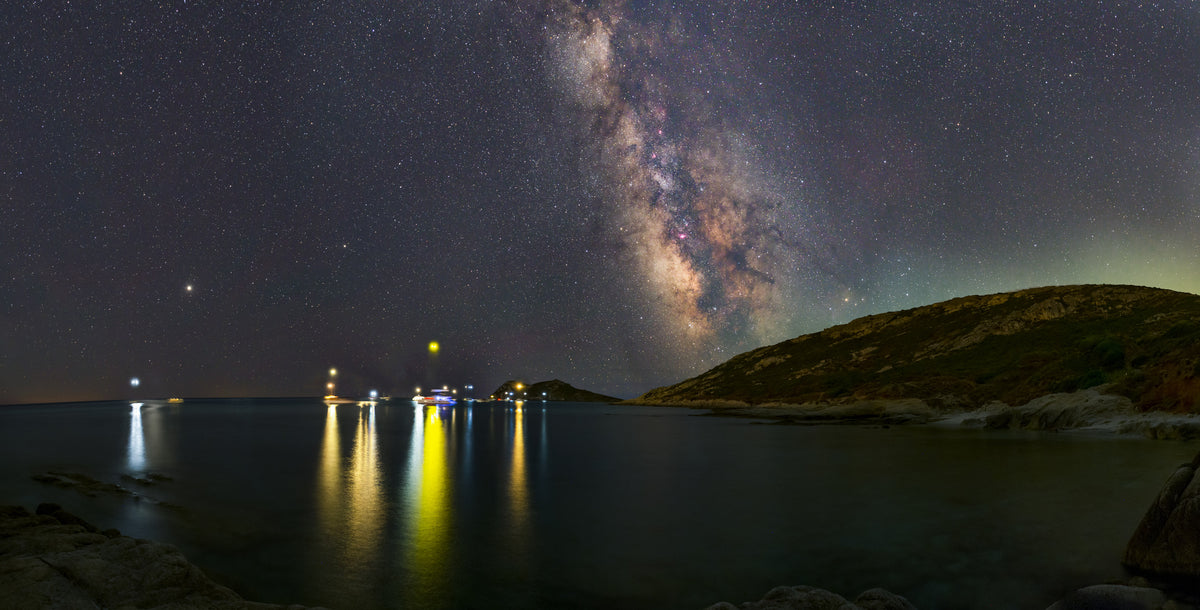 Voie lactée verticale au-dessus d’une mer sombre, avec des reflets jaunes de lumières côtières.
