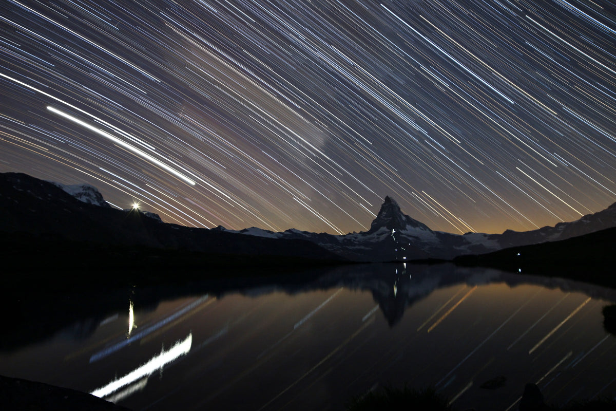 Traînées d’étoiles au-dessus du Cervin et de son reflet, avec de petites lumières sur le relief et au premier plan.