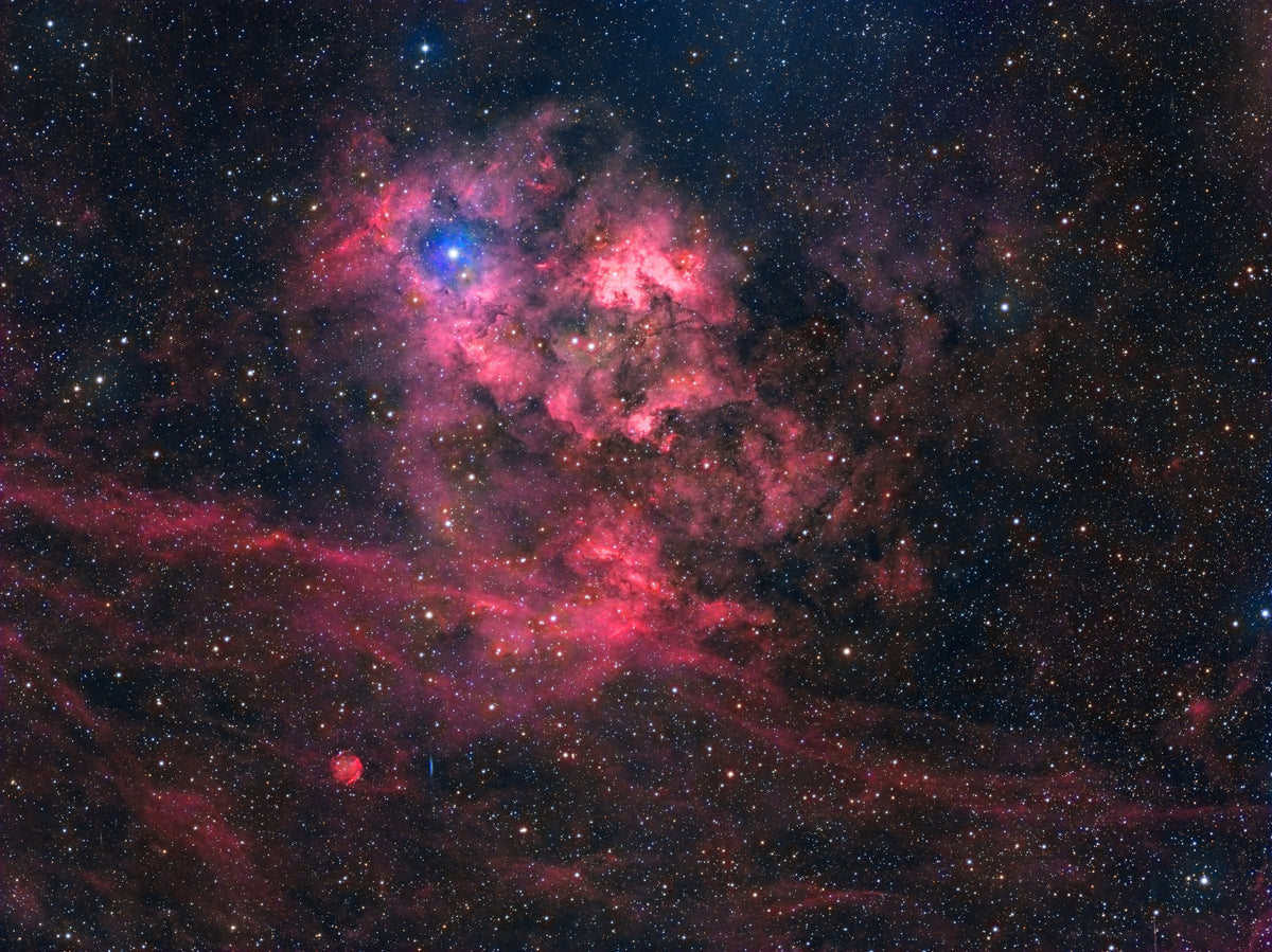Nébuleuse de la Grotte aux nuages rouges et bleutés entourant une cavité sombre, dans un champ d’étoiles.