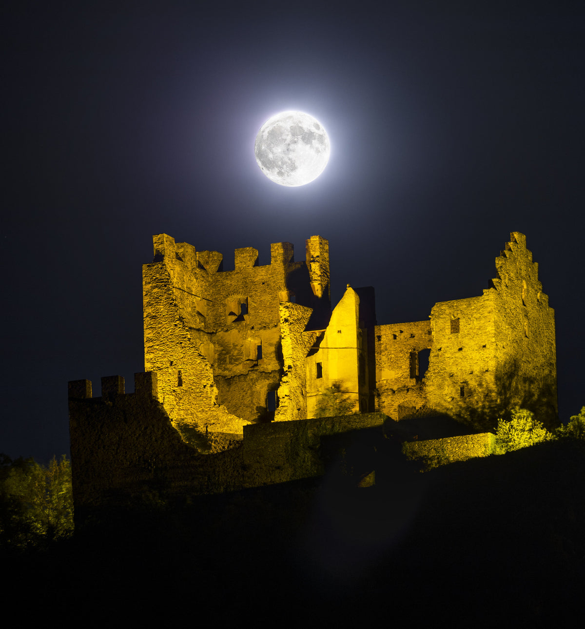 Pleine Lune au-dessus des ruines illuminées du château de Tourbillon sur une colline sombre.