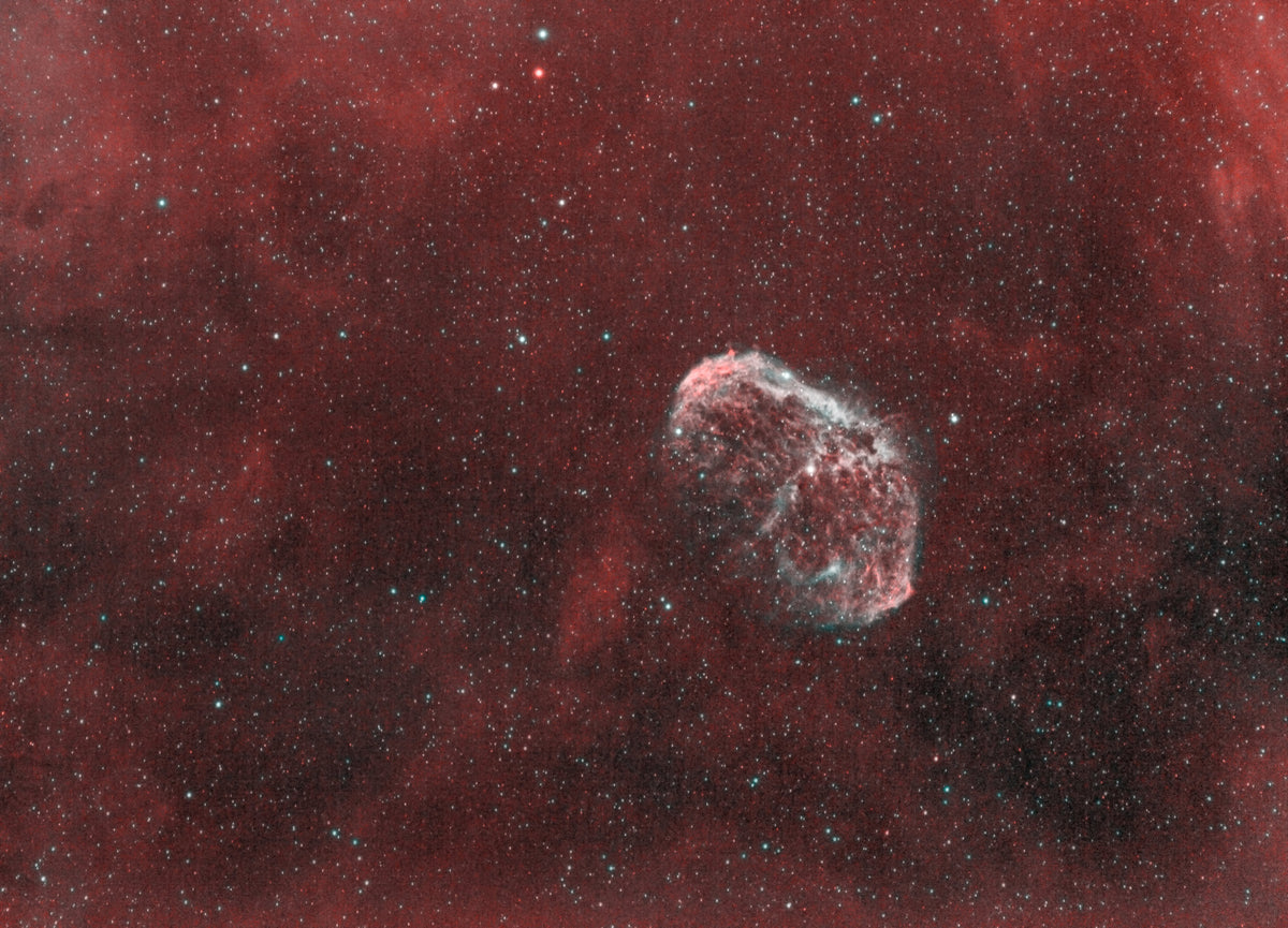 Coquille claire de la nébuleuse du Croissant, au milieu de nébulosités rouges et d’un champ d’étoiles.