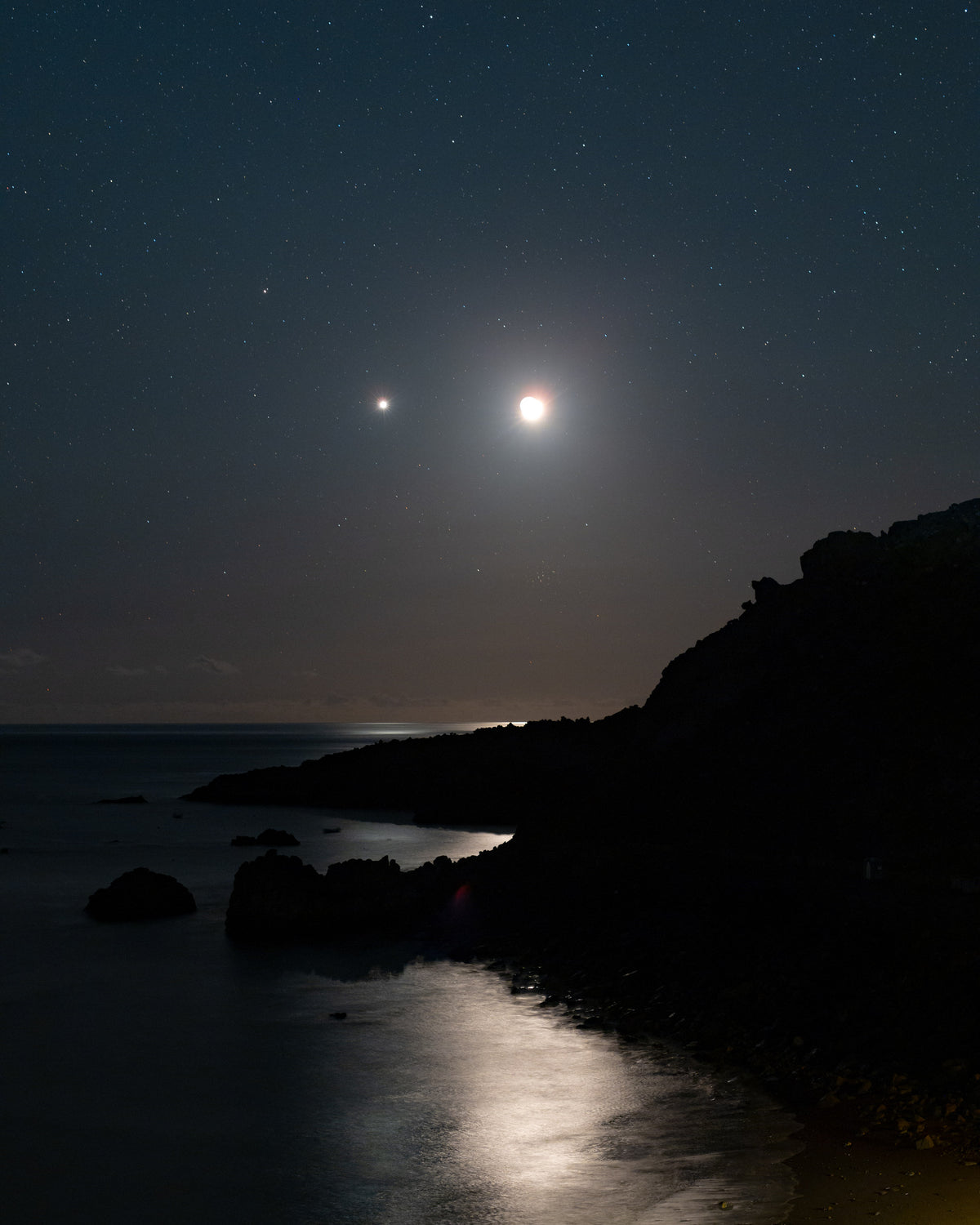 Lune et point lumineux de Vénus dans le ciel, avec un reflet clair sur l’océan et une côte en silhouette.